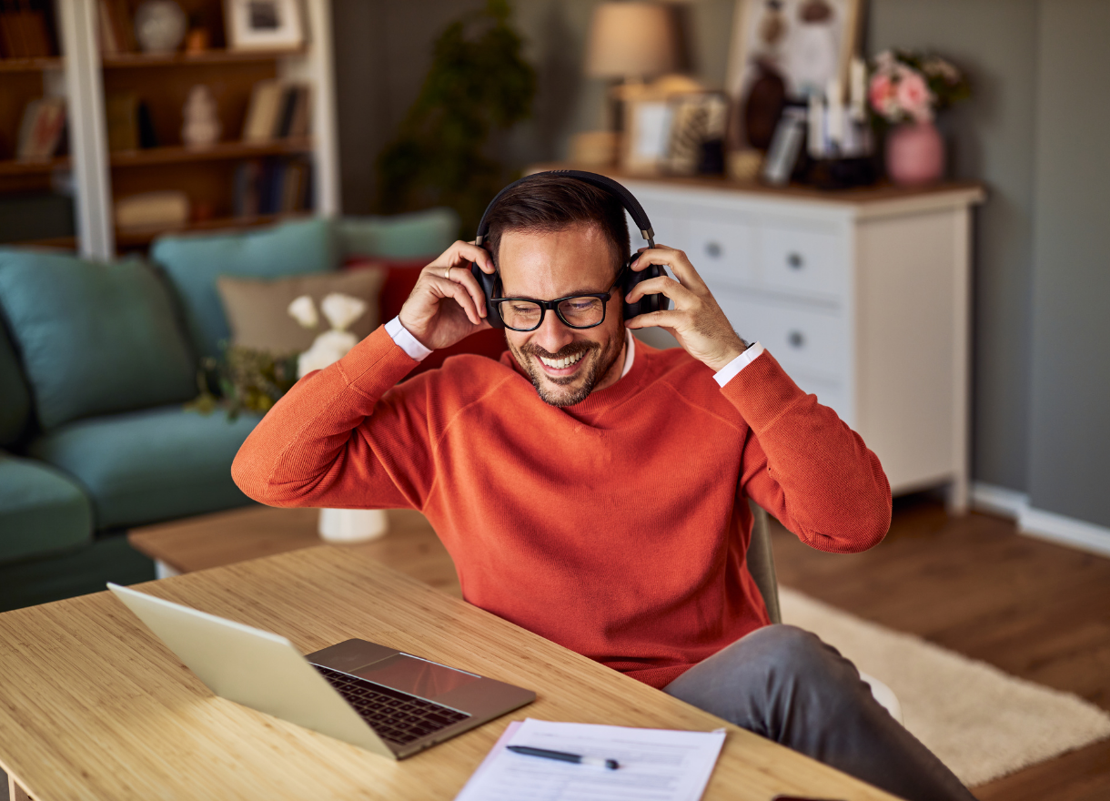 A man in an orange jumper smiling at his laptop with headphones on