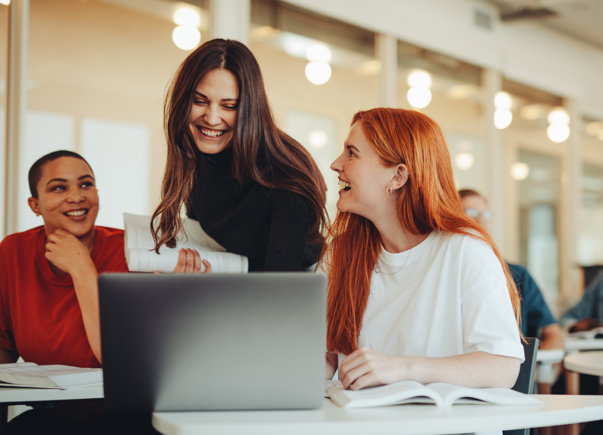 Three students laughing together over a laptop in a bright classroom